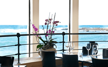 שולחן ערוך לסעודה, עציץ סחלבים וחלונות גדולים שנשקף מהם ים
