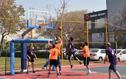 קבוצת גברים בבגדי ספורט, לחלקם חולצות בצבע אחד, ולחלקם בצבע אחר, משחקים כדורסל-רחוב במגרש כדורסל.