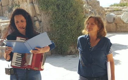אישה אוחזת קלסר ואקורדיון, מיקרופון לצד פיה, ואישה נוספת מחייכת, על רקע מבנה עתיק