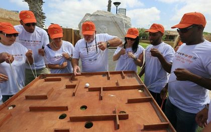 המירוץ לליגיון - פעילות גיבוש לצוותים ולחברות: קבוצת אנשים בתלבושת אחידה עומדים סביב שולחן עץ שיש בו חורים ומבוך וכדור קטן ובידיהם חוטים המאפשרים להם להטות את השולחן.