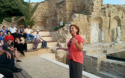 מדריכה מדברת אל קבוצת אנשים היושבת על ספסלים בקרבת עתיקות קיסריה