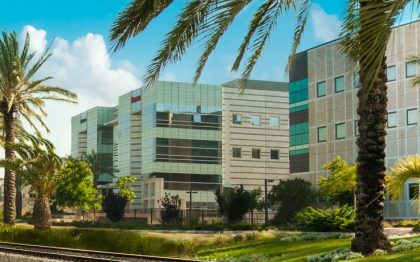 רכבת נוסעת על גבי מסילה ולצידה דקלים ומבנים עסקיים בפארק העסקים