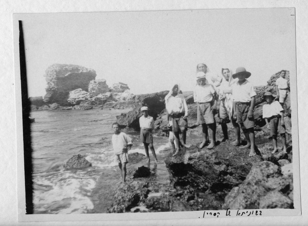תצלום בשחור-לבן של כמה דמויות חלקן גלויות ראש וחלקן חבושות כובעים, מטפחות או כפיות. הדמויות עומדות באזור סלעי סמוך לים