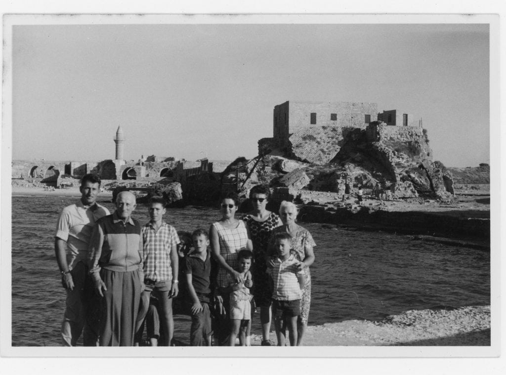 תצלום בשחור-לבן של כמה אנשים - קשישים, מבוגרים וילדים - מצטלמים יחד סמוך לים. ברקע נראים מבנים עתיקים ושרידי מבנים.