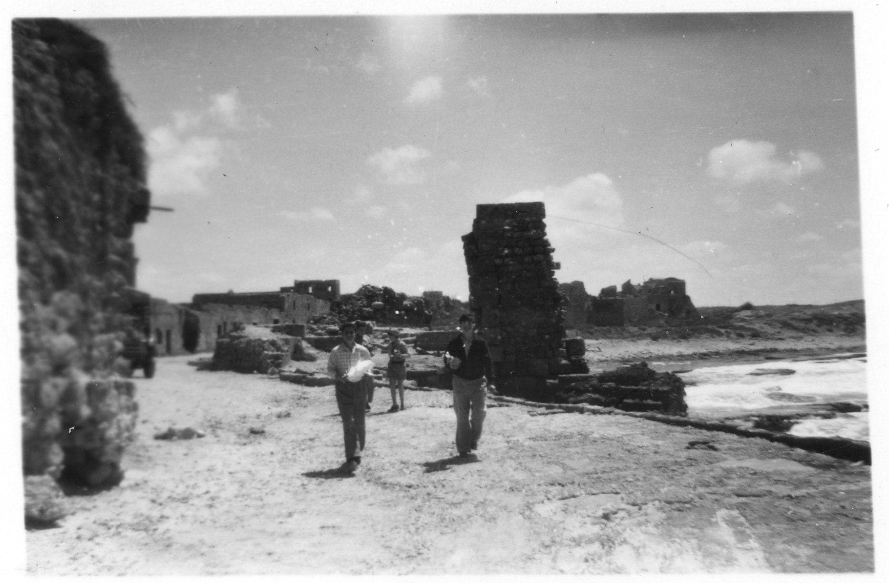 תצלום בשחור-לבן של שרידי מבנים על חוף הים. כמה נערים פוסעים על דרך לא סלולה בין המבנים.