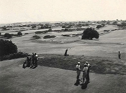 תצלום בשחור-לבן המראה שחקני גולף בבגדים של פעם, משוחחים כשבידיהם מחבטי הגולף על מגרש גולף רחב ידיים