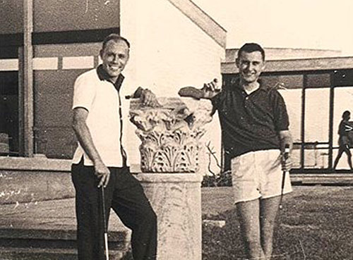 תצלום בשחור-לבן של שני בחורים אוחזים במחבטי גולף מחייכים למצלמה ומשעינים את ידיהם על חלק מכותרת עמוד עתיק מעוטר. מאחוריהם מבנה מודרני לתקופתם