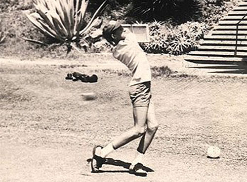 דמות בבגדים קצרים וכובע מצחייה לקראת חבטה בכדור גולף המונח על הדשא