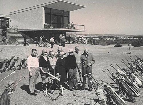 תצלום בשחור-לבן של כמה אנשים עומדים מאחורי שורה של עגלות גולף. ברקע מבנה דו קומתי שמרפסת ״תלויה״ בחזיתו.