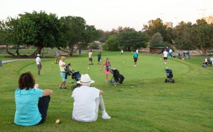 גולפאים מתאמנים במשחק הגולף במדשאה עם שיפועים קלים. בתצלום רואים גם אנשים היושבים על הדשא וכן עגלות גולף