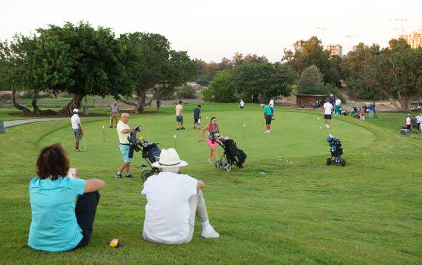 גולפאים מתאמנים במשחק הגולף במדשאה עם שיפועים קלים. בתצלום רואים גם אנשים היושבים על הדשא וכן עגלות גולף