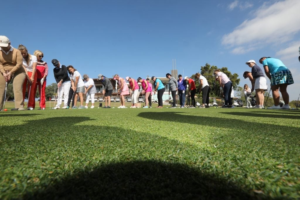 גברים ונשים עומדים על הדשא זה אחר זה בחצי גורן ואוחזים במחבטי גולף וחובטים בכדורי גולף למרכז המעגל.