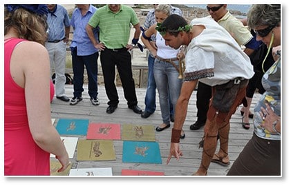 מדריך לבוש ברוח קיסריה העתיקה מציג לקבוצת משתתפים ציורים צבעוניים המונחים על רצפת עץ חיצונית.