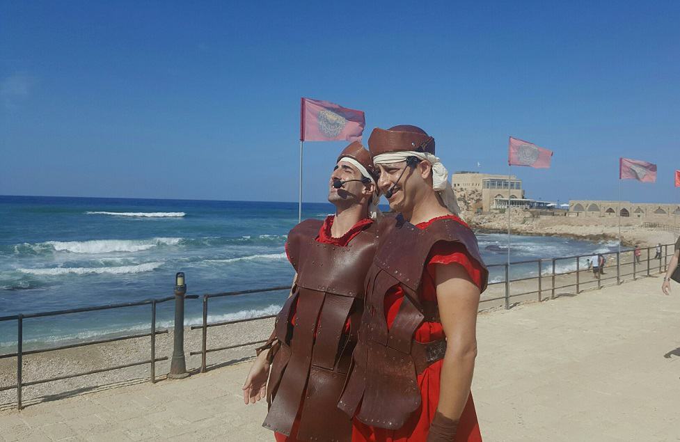 שחקנים לבושים בתלבושת תקופתית המדמה בין השאר שריון אבירים, עומדים על חוף הים בנמל קיסריה.