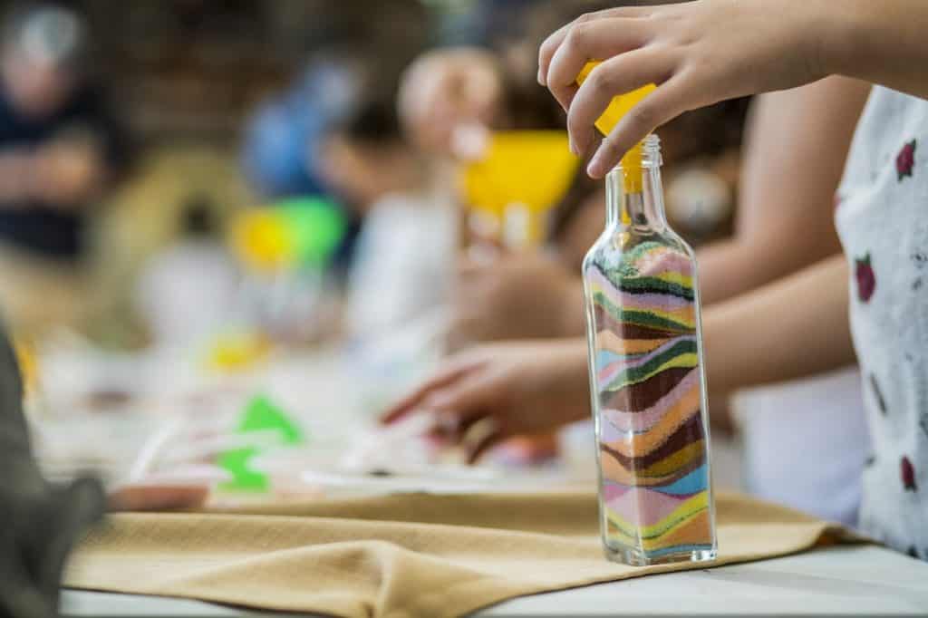 בקבוק שקוף ובו שכבות של חול צבעוני מונח על שולחן יצירה ואחוז בידי נערה.