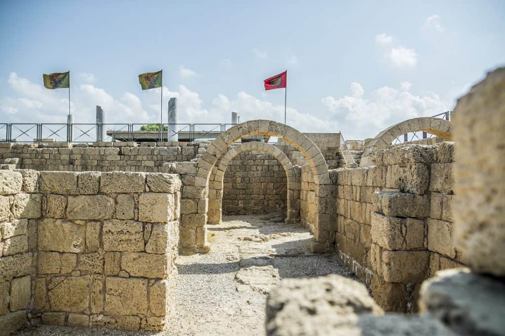 תצלום בגן לאומי קיסריה: קירות אבן וקשתות אבן. מרחוק נראים שלושה דגלים