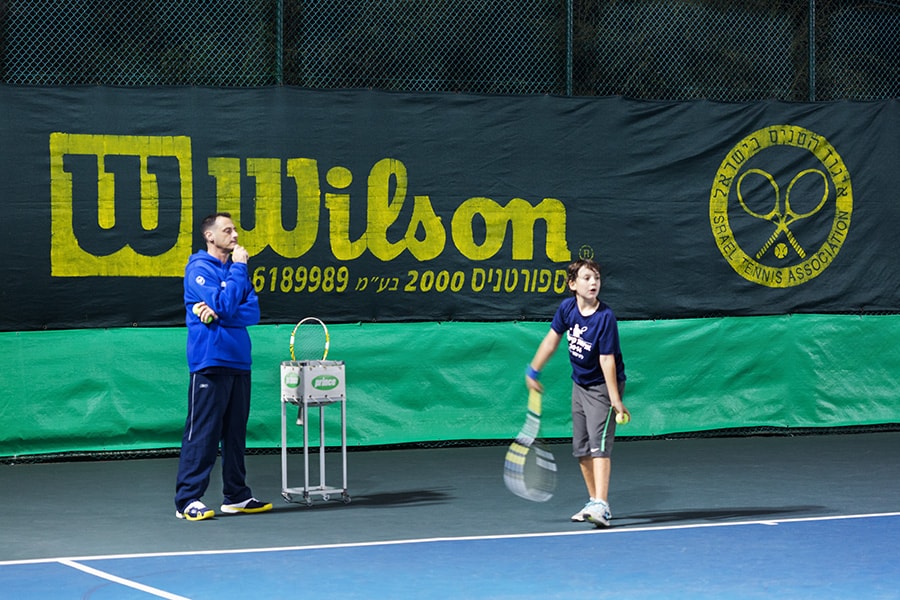 נער נערך לחבטת הגשה בטניס. לצידו מאמן מביט במהלך.