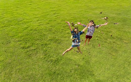 שתי ילדות מזנקות מעל כר דשא, בזרועות פתוחות, וסביבן מעופפים עלים יבשים