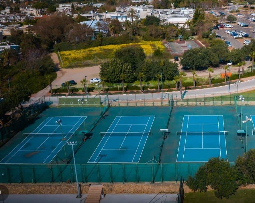 תצלום מן האוויר של שלושה מגרשי טניס צמודים, אזור חניה, שטח ירוק וסביבת מגורים.