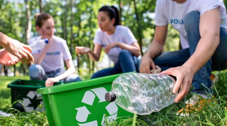 צעירים ממחזרים בקבוקי פלסטיק במיכלים ייעודיים בשטח ירוק של דשא ועצים