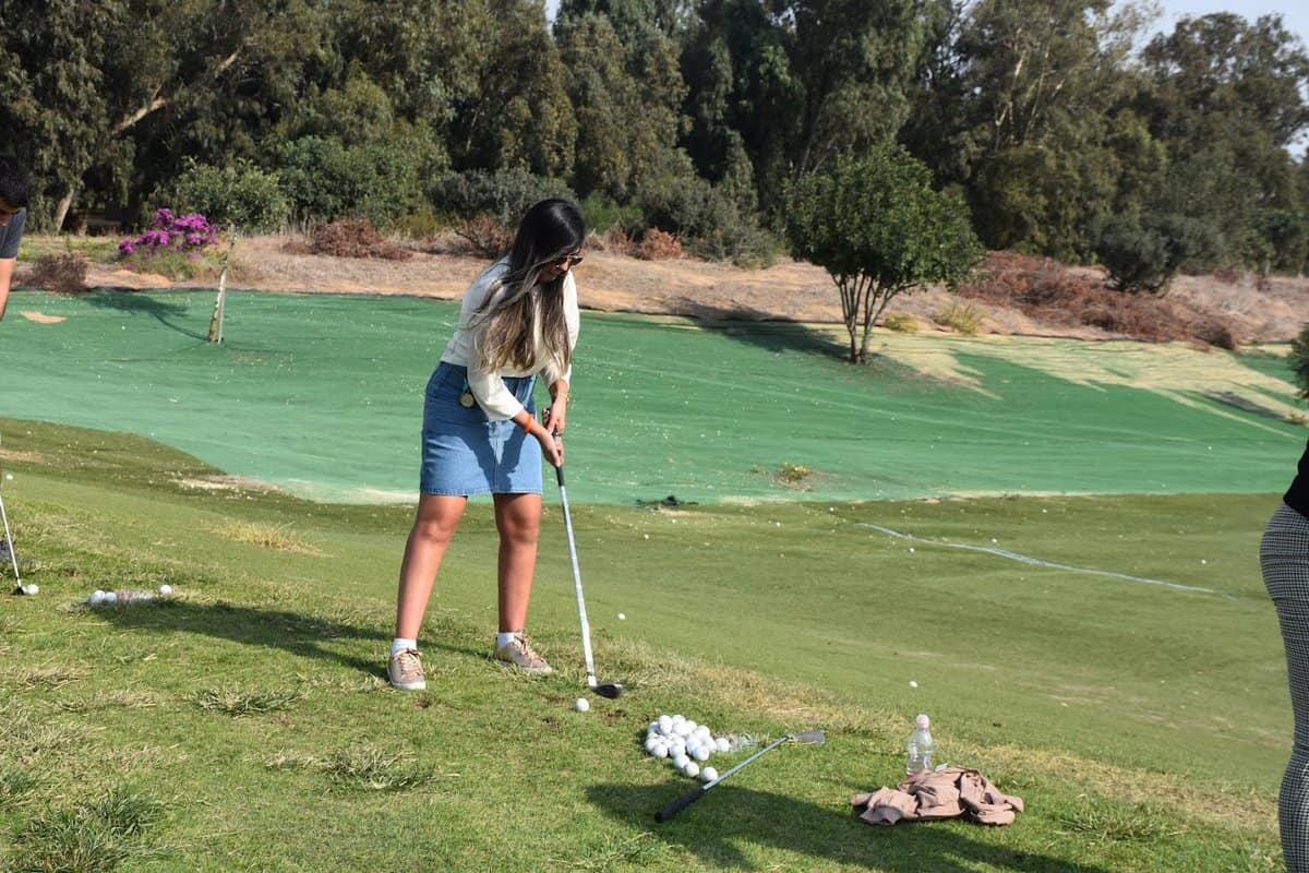 בחורה אוחזת במחבט גולף לקראת חבטה בכדור גולף. לידה מקבץ של כדורים נוספים. הדשא שעליו היא עומדת נמצא בשיפוע