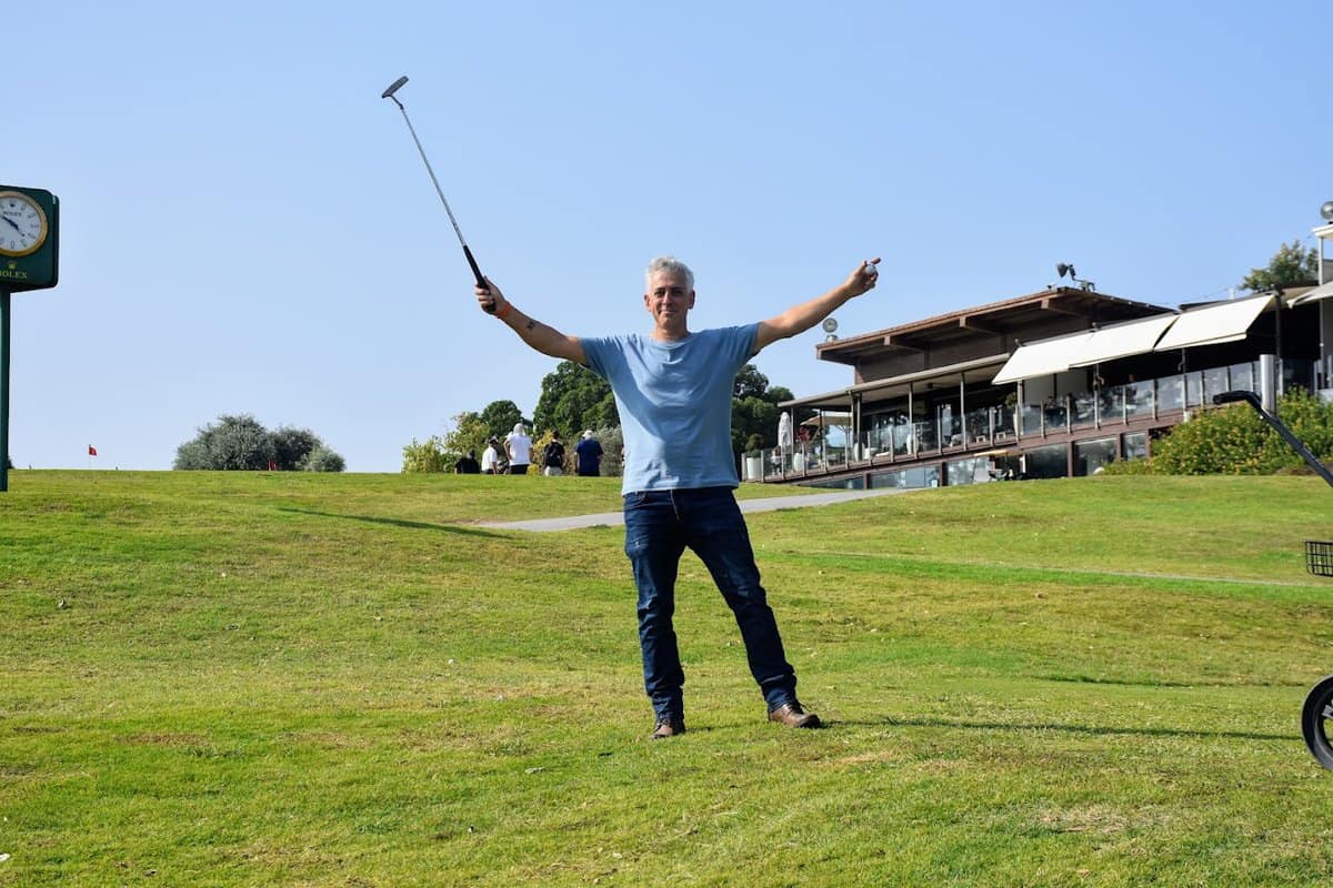 גבר אוחז במחבט גולף מונף כלפי מעלה, שתי ידיו פתוחות לצדדים, ומאחוריו כרי דשא ומבנה