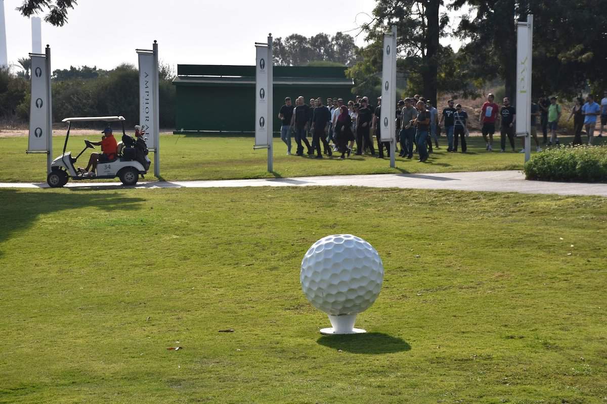 קבוצת אנשים פוסעת במגרש גולף בעקבות מישהו שנוהג ברכב גולף. כדור גולף גדול מונח על כן בחזית התמונה