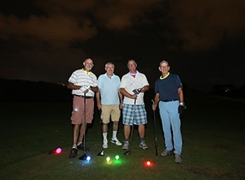 ארבעה גברים עם מחבטי גולף עומדים בשורה בלילה ולפניהם תאורה צבעונית על הדשא