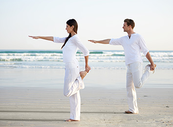 אישה וגבר בבגדים לבנים על חוף הים מושיטים יד אחת קדימה, ויד אחרת אוחזת בכף הרגל מאחור